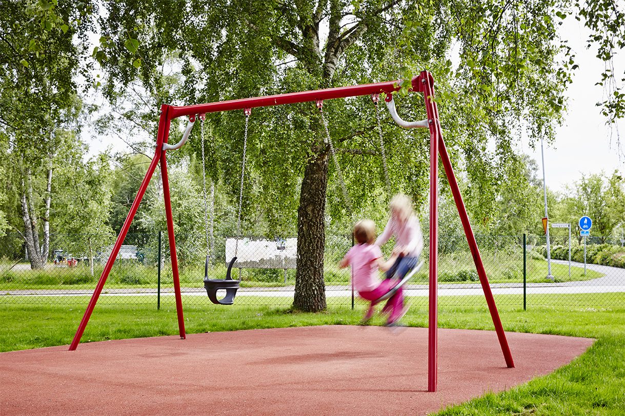 Children swinging on a red steel swing frame in a small playground next to a road